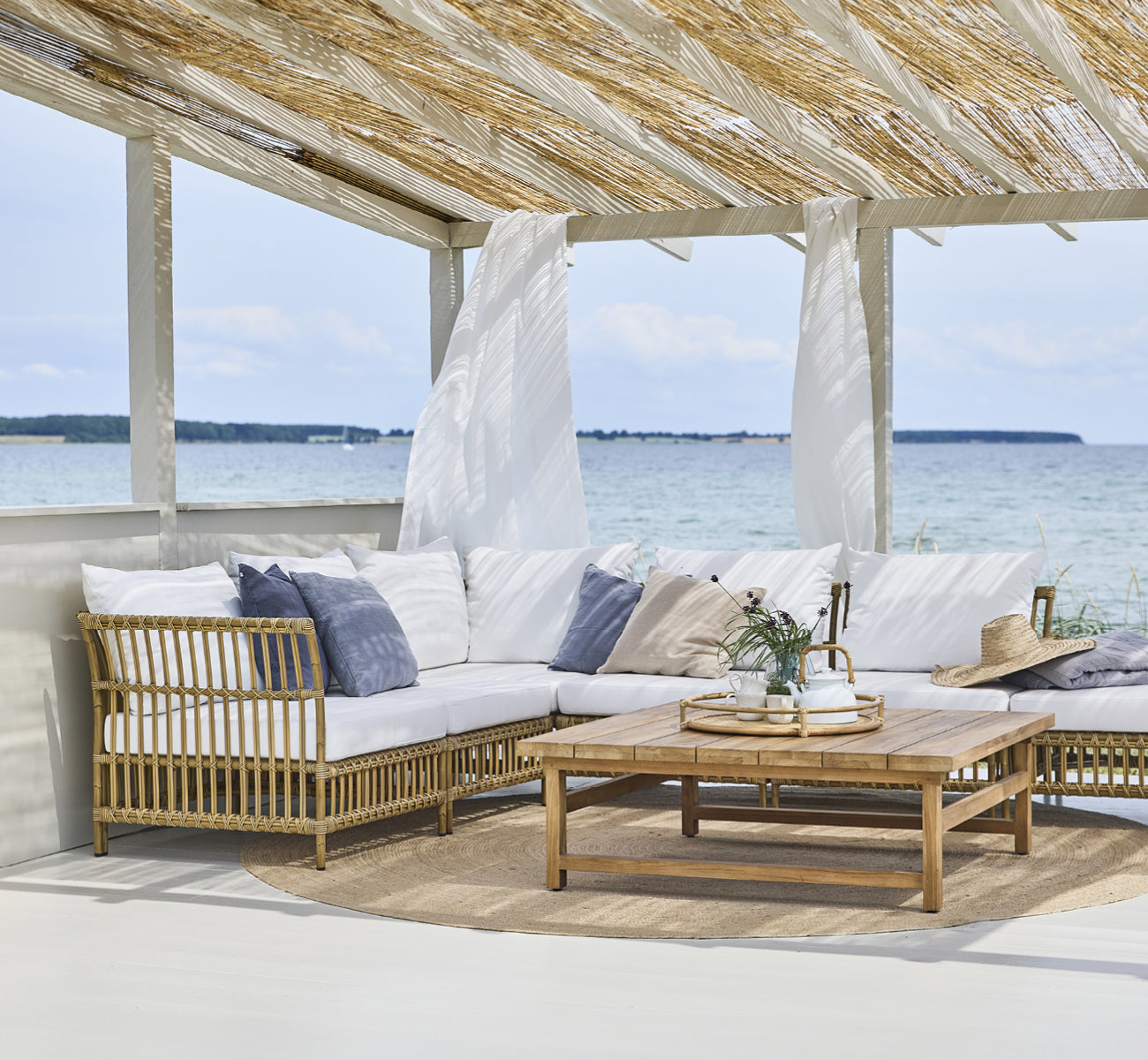 Esta es una imagen de estilo de vida de Julian Teak Lounge Table 120x120 cm en teak outdoor. Bajo una pérgola de playa con asientos lounge y vistas al océano.