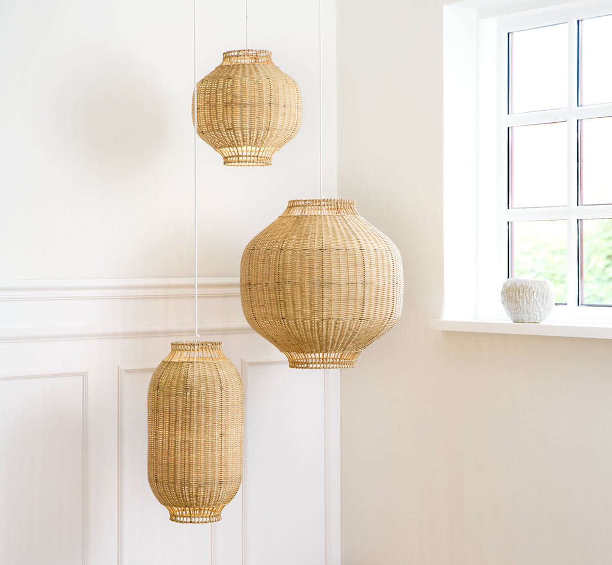 Esta es una imagen de estilo de vida de Tomos pantalla de lámpara en natural core weave rattan frame. Colgada con otros tamaños de colgante en una luminosa sala con paneles.