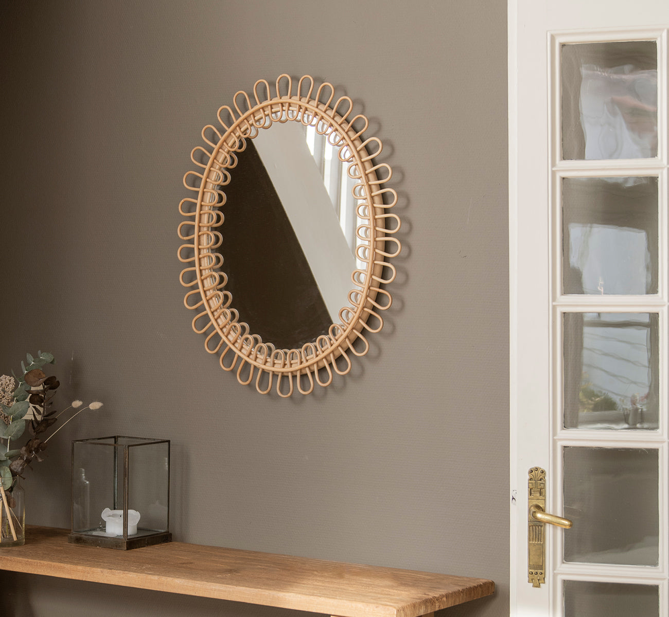 Esta es una imagen de estilo de vida de Luella espejo en polished natural rattan. Colgado en una pared gris sobre una estantería de madera con eucalipto y decoración de cristal.