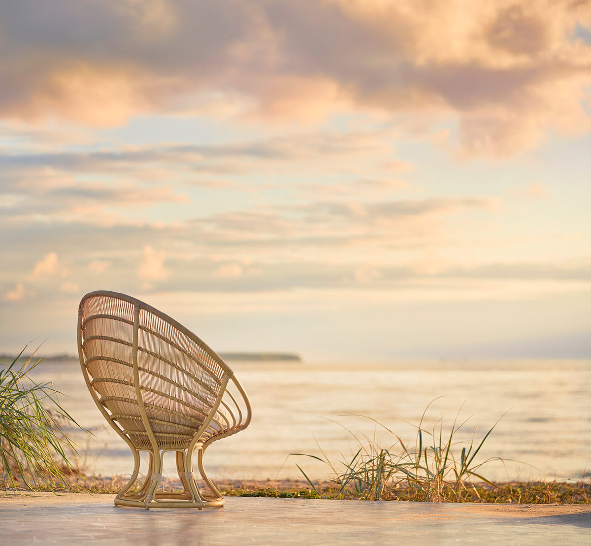 Esta es una imagen de estilo de vida de Luna sillón lounge exterior en natural aluminium artfibre. Vista desde atrás en una playa al atardecer con un cielo de tonos pastel.
