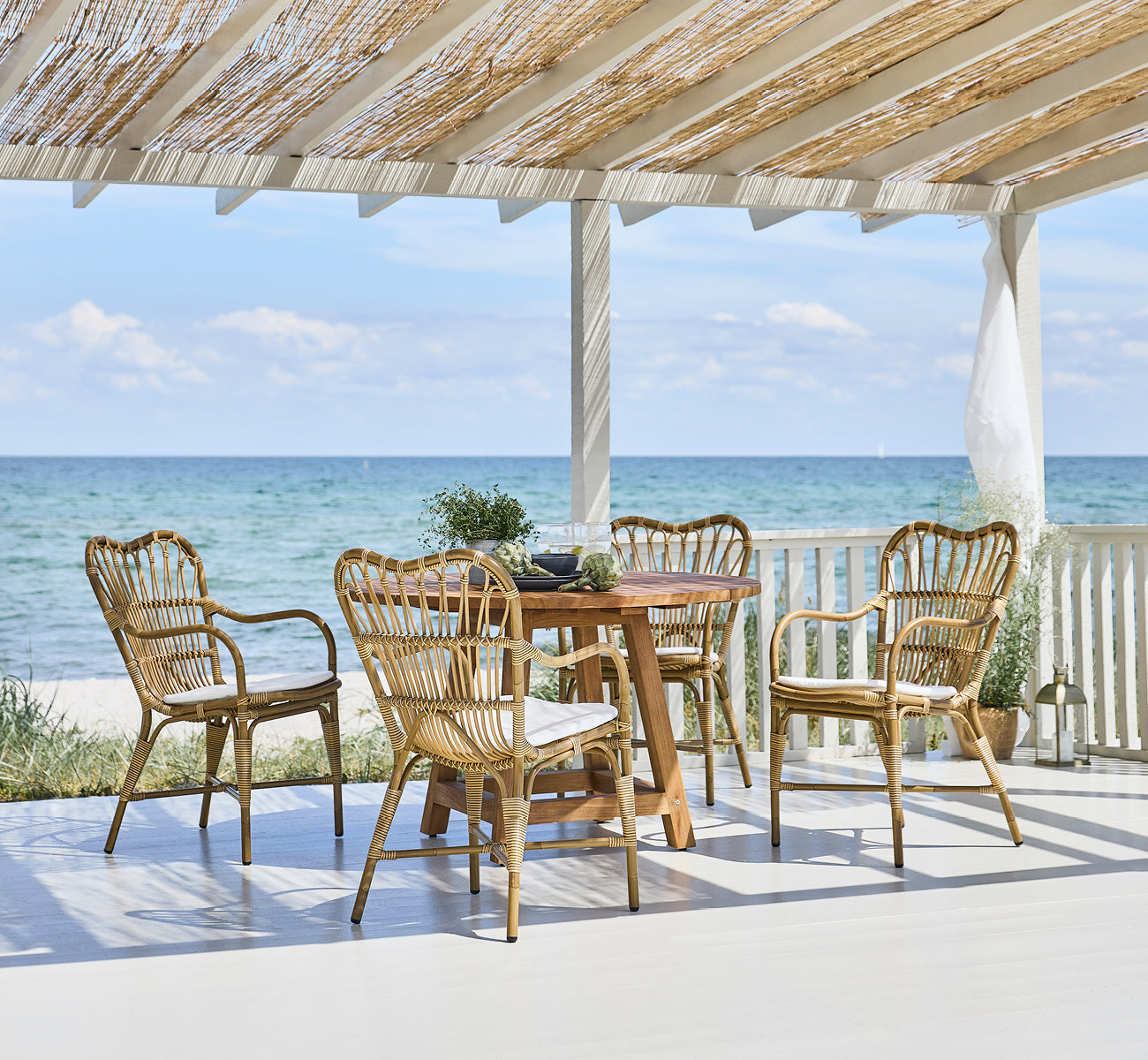 Esta es una imagen de estilo de vida de Margret sillón de comedor exterior en antique aluminium artfibre. Pabellón de playa con mesa de teca y vista al mar.