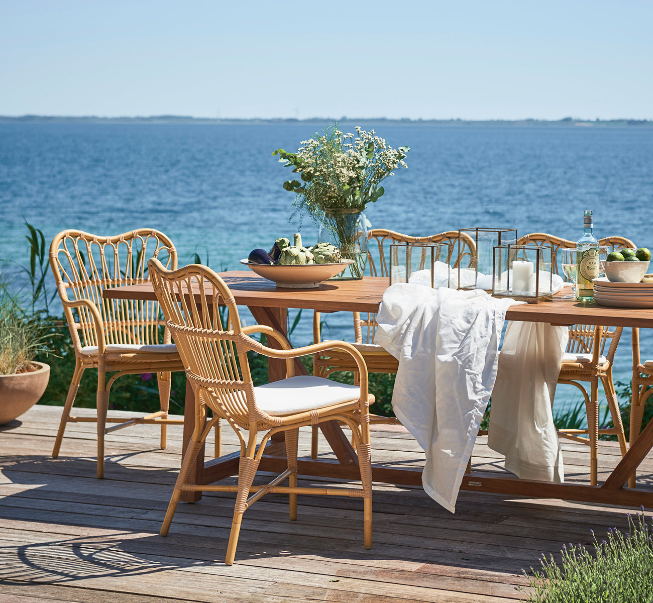 Esta es una imagen de estilo de vida de Margret sillón de comedor exterior en natural aluminium artfibre. Terraza de madera junto al mar con mesa de teca y flores.