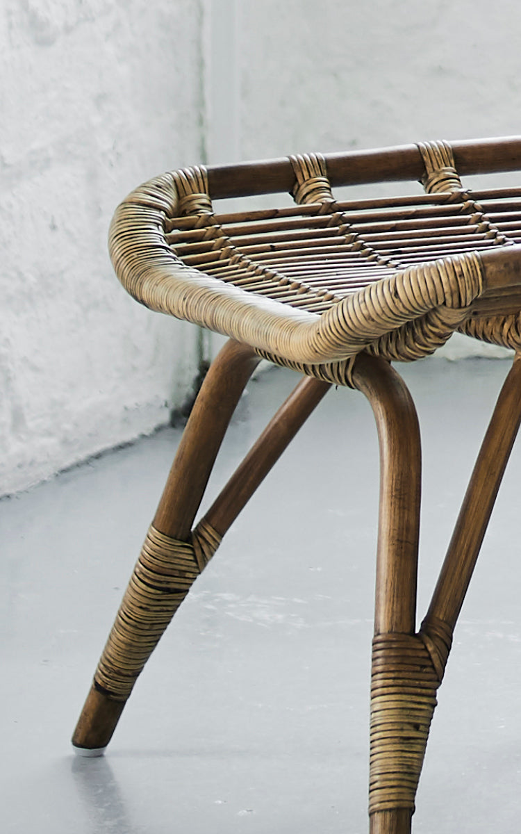 Esta es una imagen de detalle de Monet Footstool i antique rattan. Detalle de la superficie tejida plana del asiento y las patas abiertas de ratán.