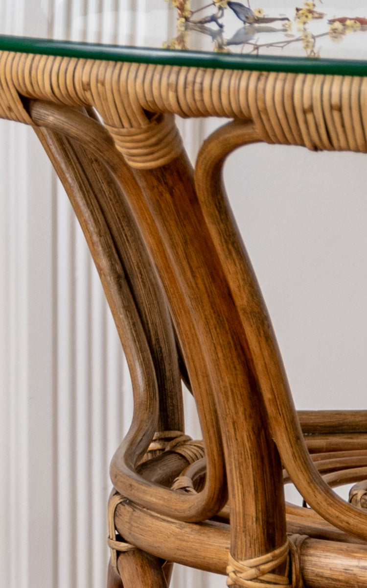 Esta es una imagen de detalle de Tony Café Table en antique rattan. Detalle del borde de la tapa de vidrio, estructura curva de ratán y uniones envueltas.