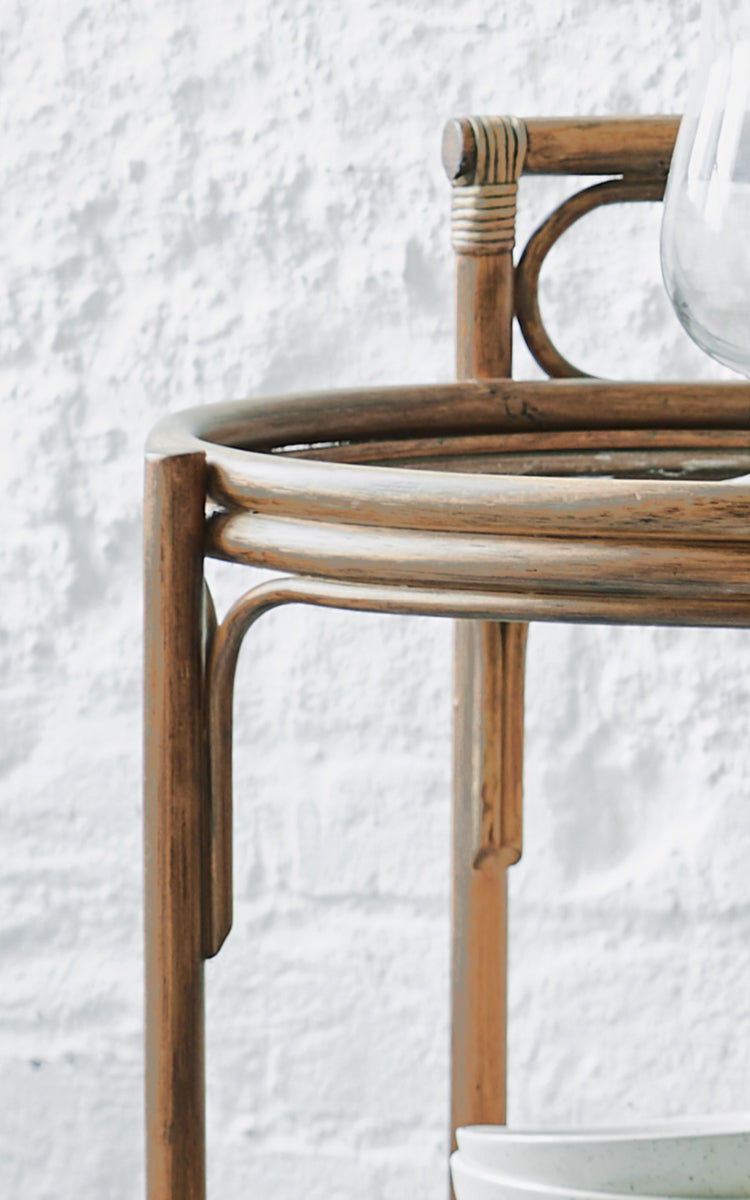 Esta es una imagen de detalle de Romeo carrito en antique rattan. Detalle de la estructura redonda del carrito de ratán sobre una pared de ladrillo blanco.
