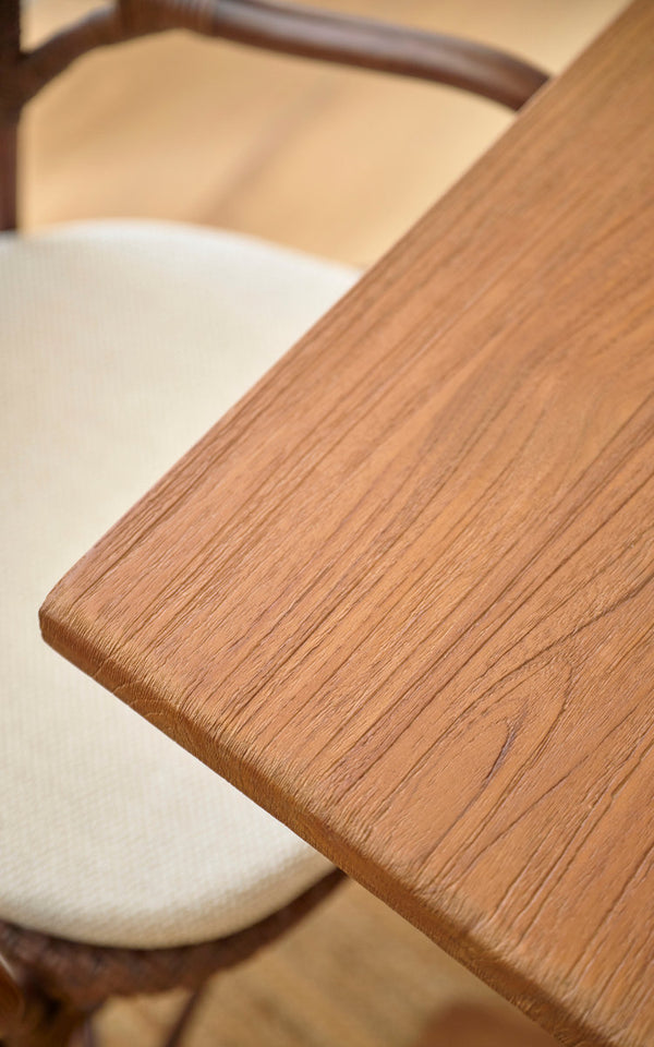 Esta es una imagen de detalle de Hugo mesa de comedor de teca 140x90 cm en teak indoor. Borde liso de la tapa de teca junto a una silla de ratán.