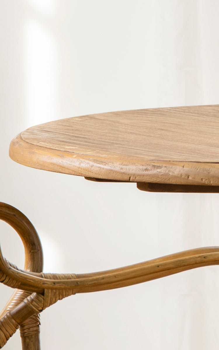 Esta es una imagen de detalle de Michel Teak Dining Table Ø80 cm en teak indoor. El borde redondo de la mesa y la base curva de pedestal de ratán se muestran.