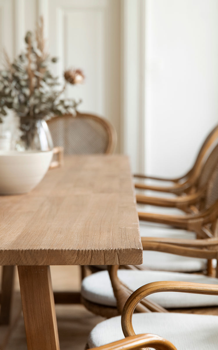 Esta es una imagen de detalle de Lucas Teak Dining Table 300X100 cm en teak indoor. El borde de teca maciza se muestra con sillas de ratán al fondo.