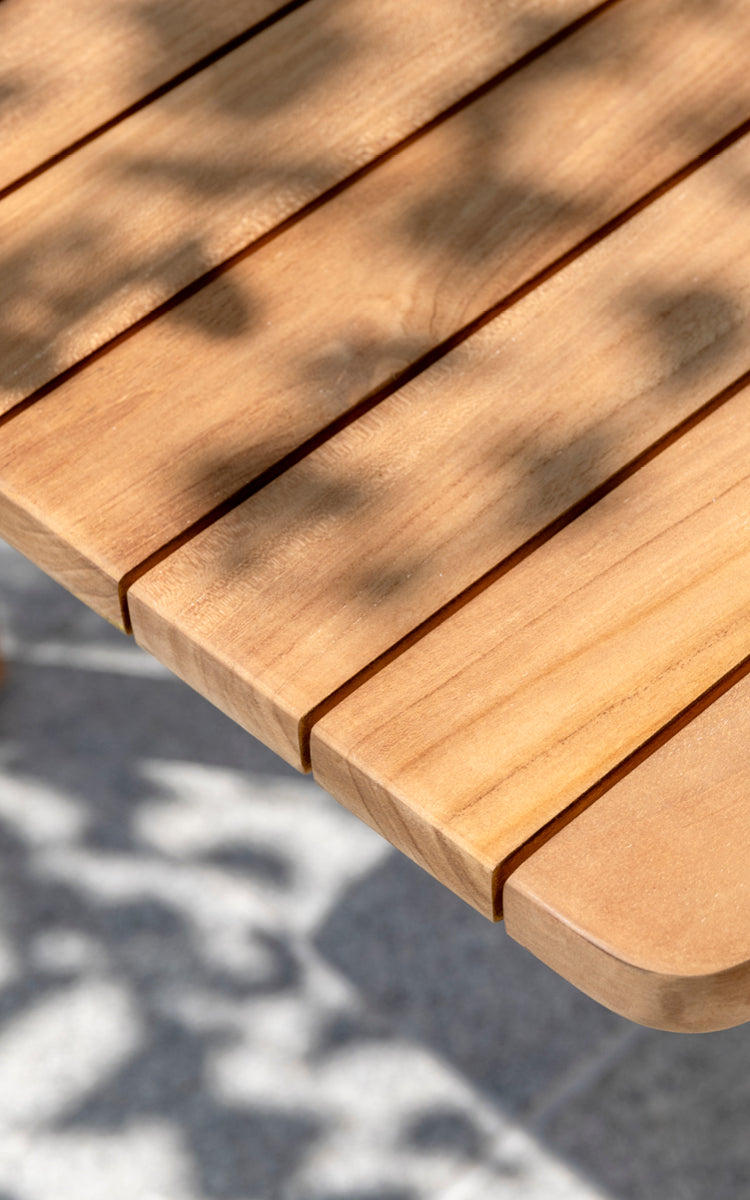 Esta es una imagen de detalle de Tobias Exterior Teak Table 205x91 cm en teak outdoor. Detalle del borde del tablero de tablones de teca y luz del sol moteada.