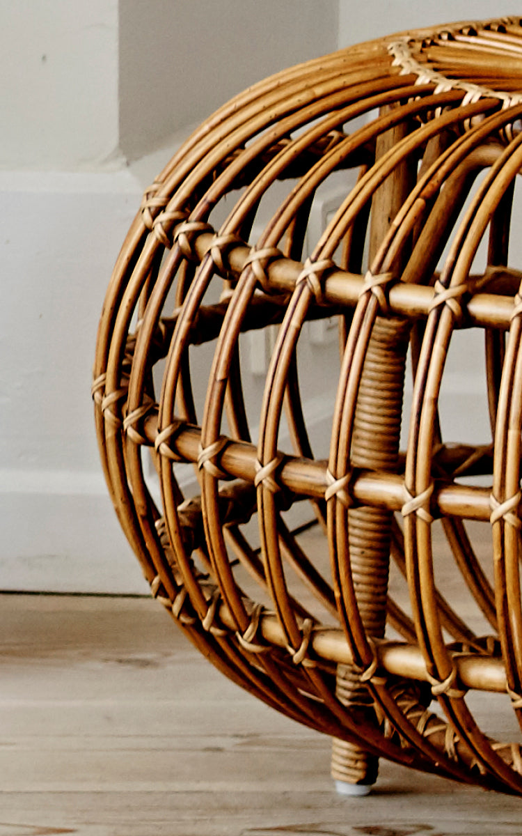 Esta es una imagen de detalle de Ottoman Large en polished antique rattan. Detalle de la gran jaula de ratán abierta con costillas curvas sobre suelo de madera.