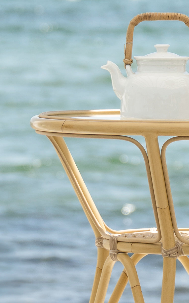 Esta es una imagen de detalle de Tony mesa de café exterior en natural aluminium artfibre. Una tetera blanca reposa sobre la superficie redonda con el mar detrás.