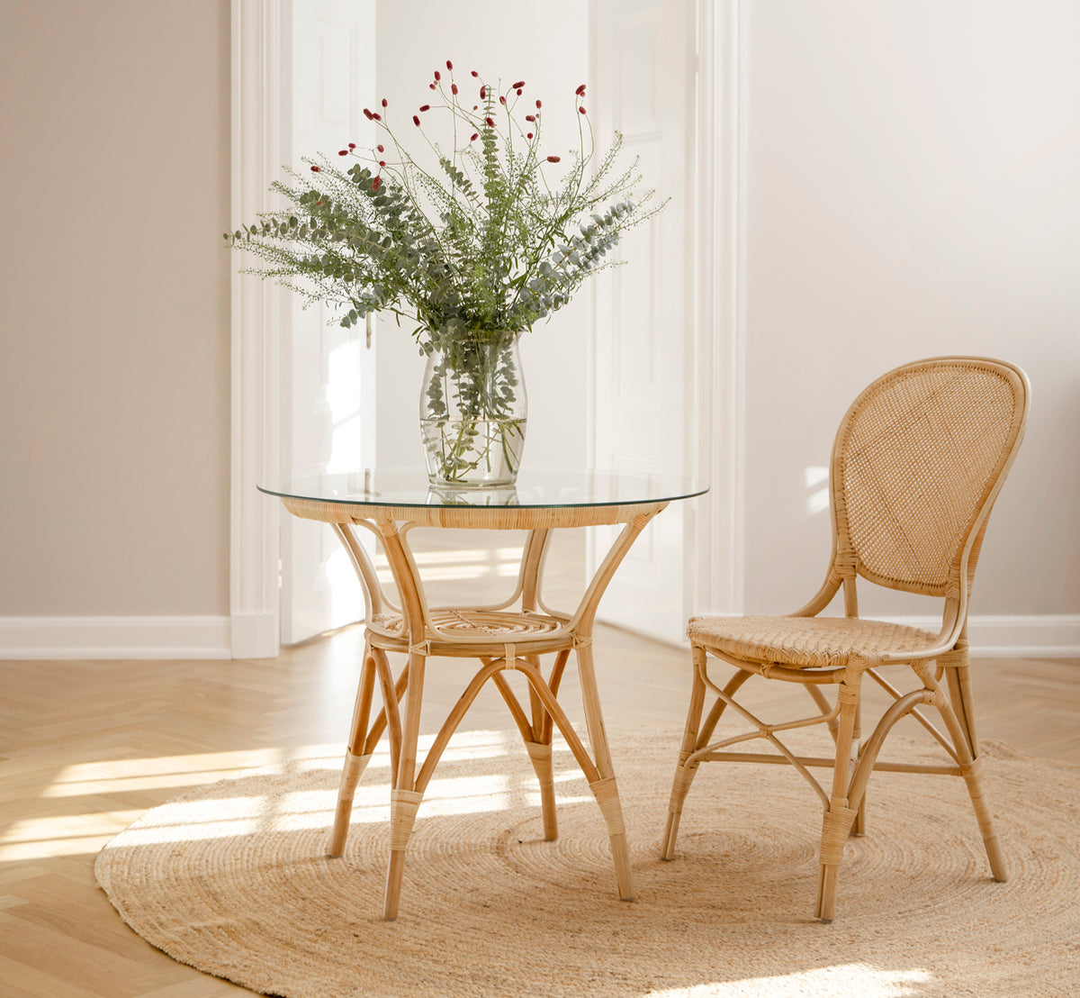 Esta es una imagen de estilo de vida de Rossini silla de comedor en natural rattan. Colocada junto a una mesa bistró de vidrio sobre una alfombra de yute en un pasillo luminoso.