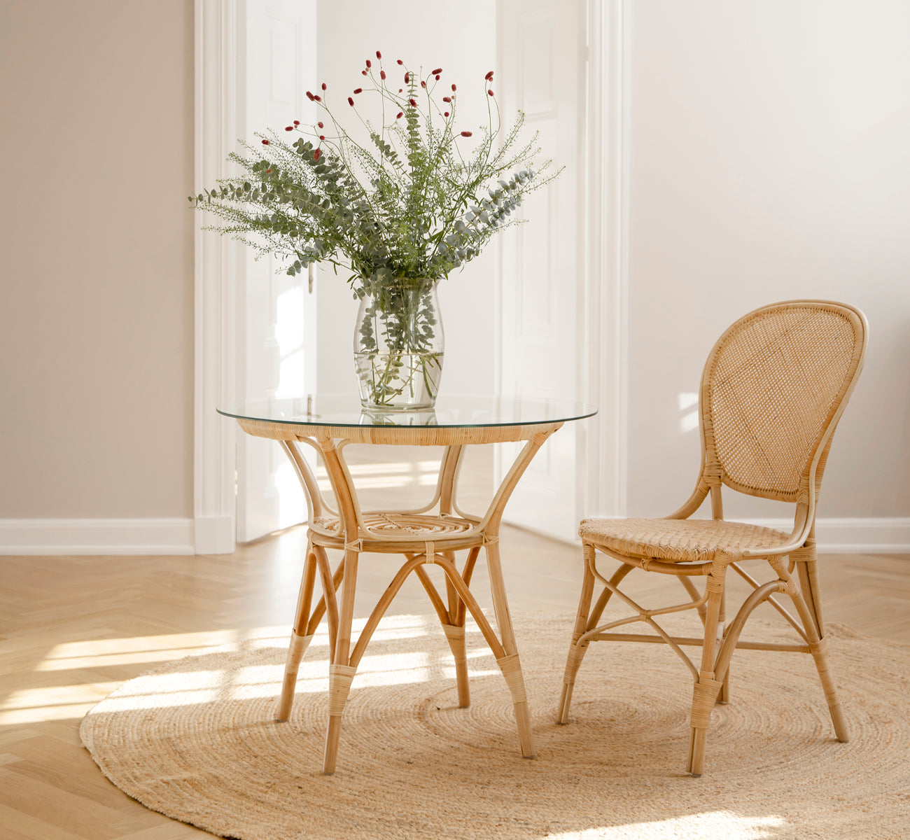 Esta es una imagen de estilo de vida de Rossini silla de comedor en natural rattan. Colocada junto a una mesa bistró de vidrio sobre una alfombra de yute en un pasillo luminoso.