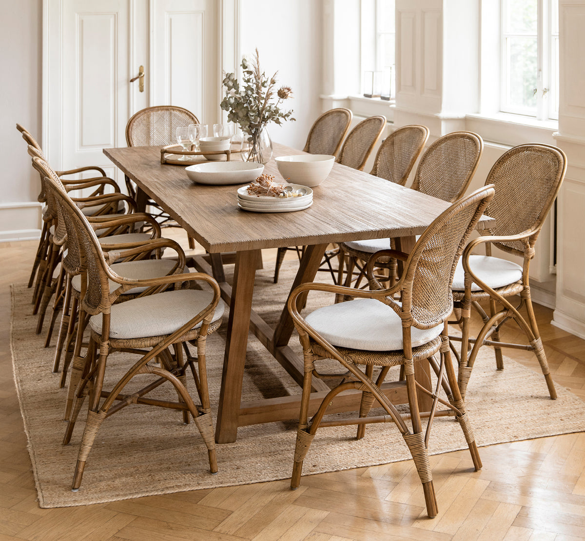Esta es una imagen de estilo de vida de Rossini sillón de comedor en antique rattan. Rodeando una larga mesa de teca con lino drapeado sobre una alfombra de yute redonda.