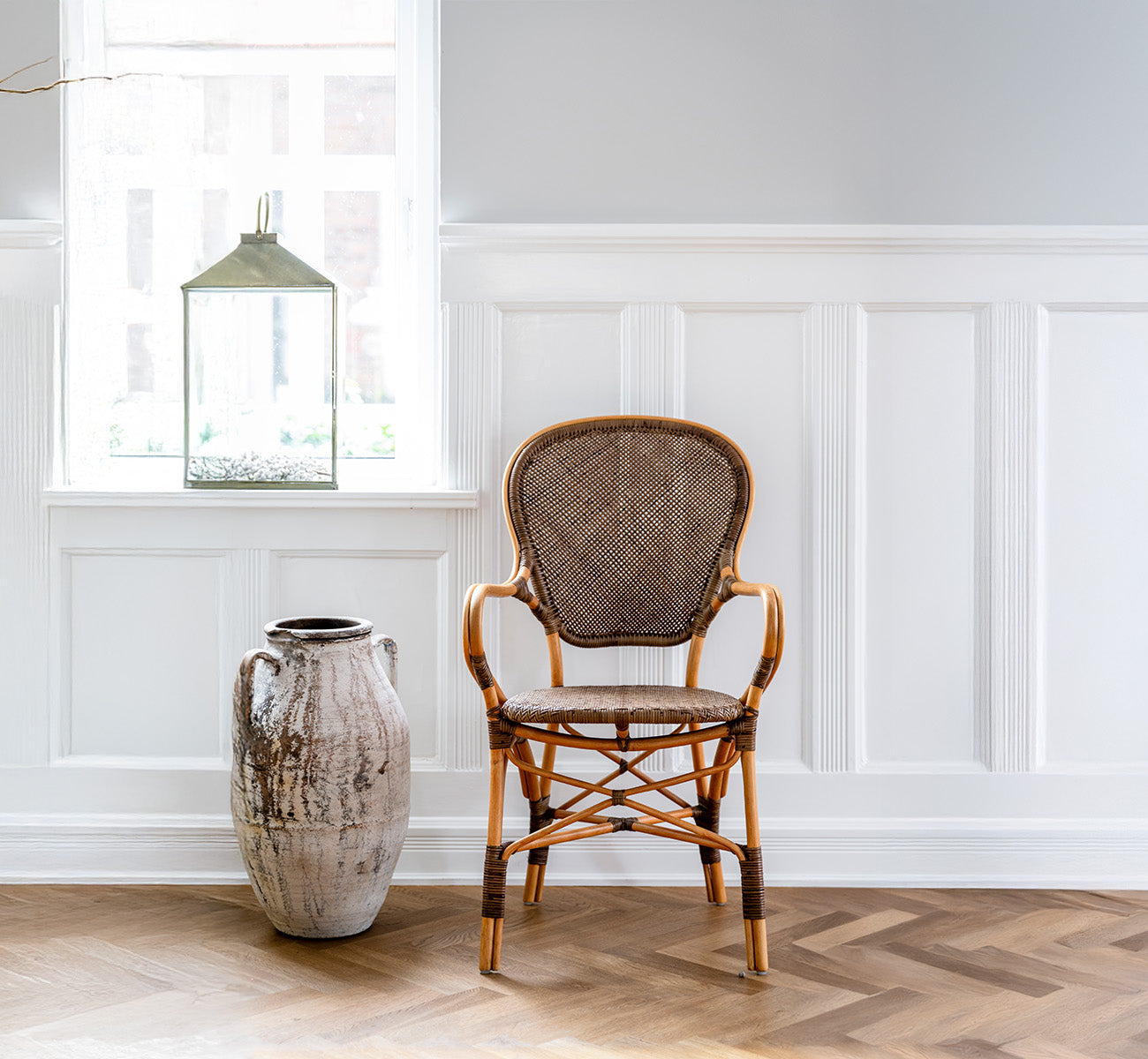 Esta es una imagen de estilo de vida de Rossini sillón de comedor en cherry rattan. Colocada en una sala con paneles y suelo en espiga junto a una urna rústica.
