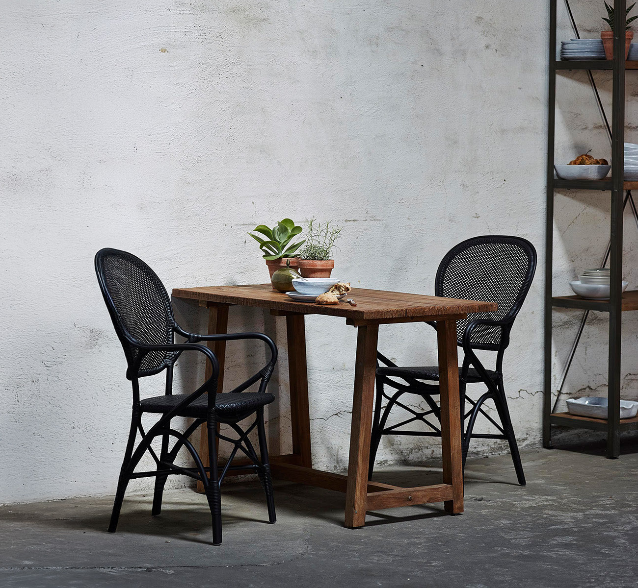 Esta es una imagen de estilo de vida de Rossini sillón de comedor en black rattan. Colocada alrededor de una mesa de madera reciclada en un loft de hormigón visto.