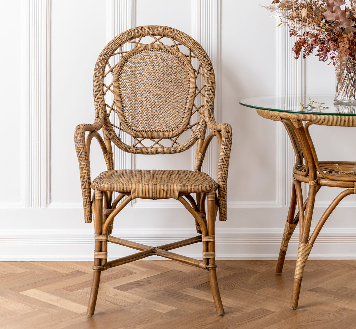 Esta es una imagen de estilo de vida de Romantica silla de comedor en antique rattan. Colocada junto a una mesa de cristal y ratán con flores secas en una sala con paneles.