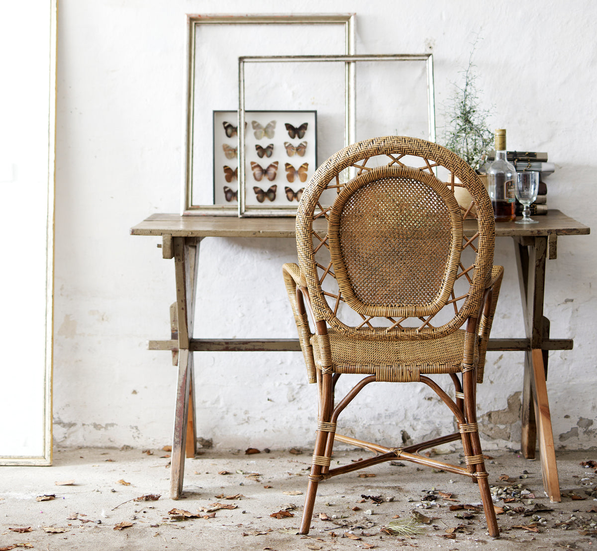 Esta es una imagen de estilo de vida de Romantica silla de comedor en antique rattan. Estilizada junto a una mesa de madera rústica con arte enmarcado en un ambiente vintage.