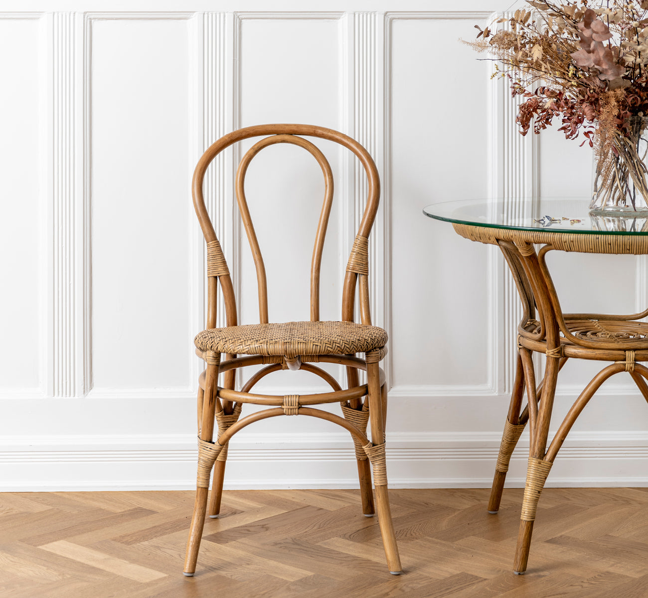 Esta es una imagen de estilo de vida de Lulu silla de comedor en antique rattan. Colocada junto a una mesa de cristal y ratán con flores secas en un interior con paneles.