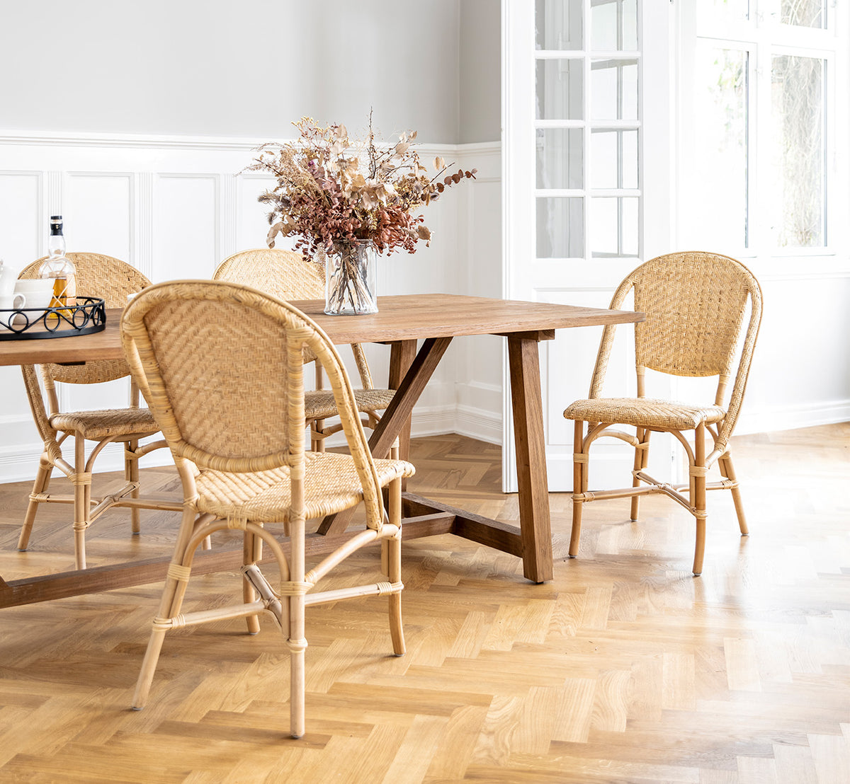 Esta es una imagen de estilo de vida de Alanis silla de comedor en natural rattan, en un interior luminoso con paredes de paneles blancos y parqué en.