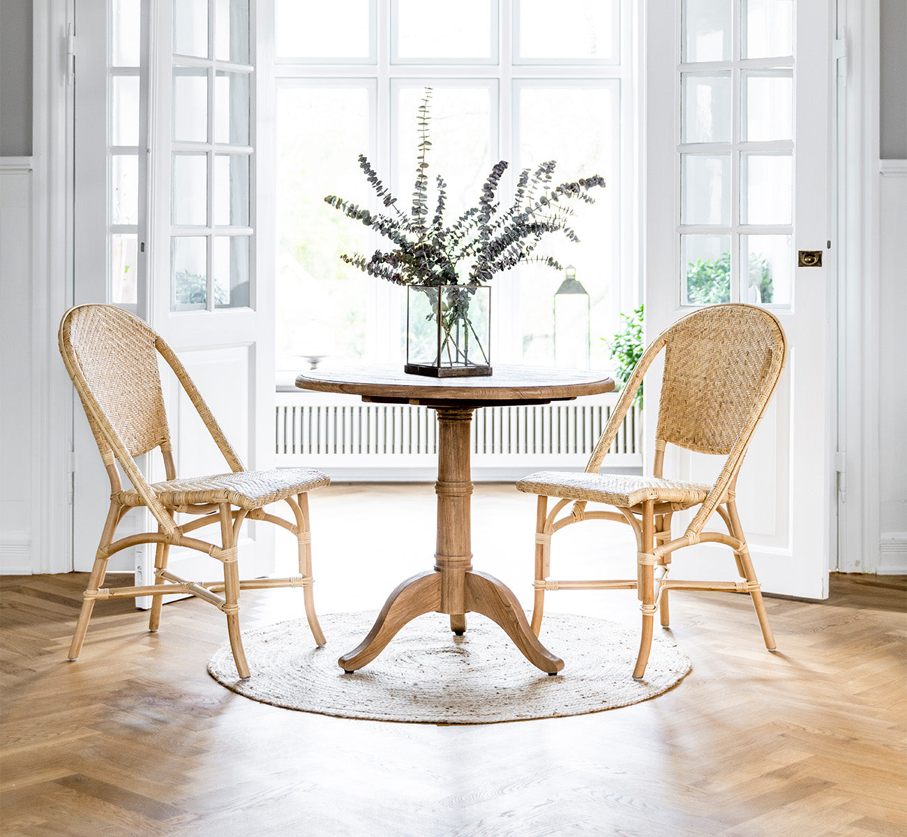Esta es una imagen de estilo de vida de Alanis silla de comedor en natural rattan, en un interior luminoso con paredes de paneles blancos y parqué en.