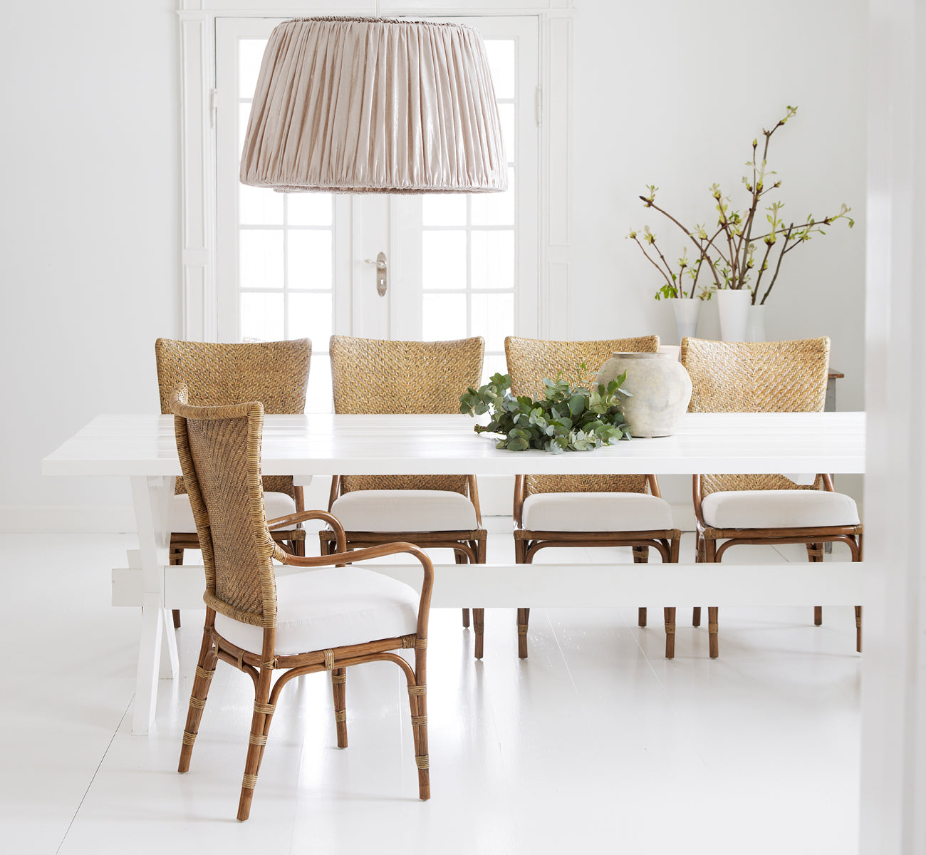 Esta es una imagen de estilo de vida de Melody Dining Armchair i antique rattan. Alrededor de una mesa blanca con lámpara colgante de lino y ramas de primavera.