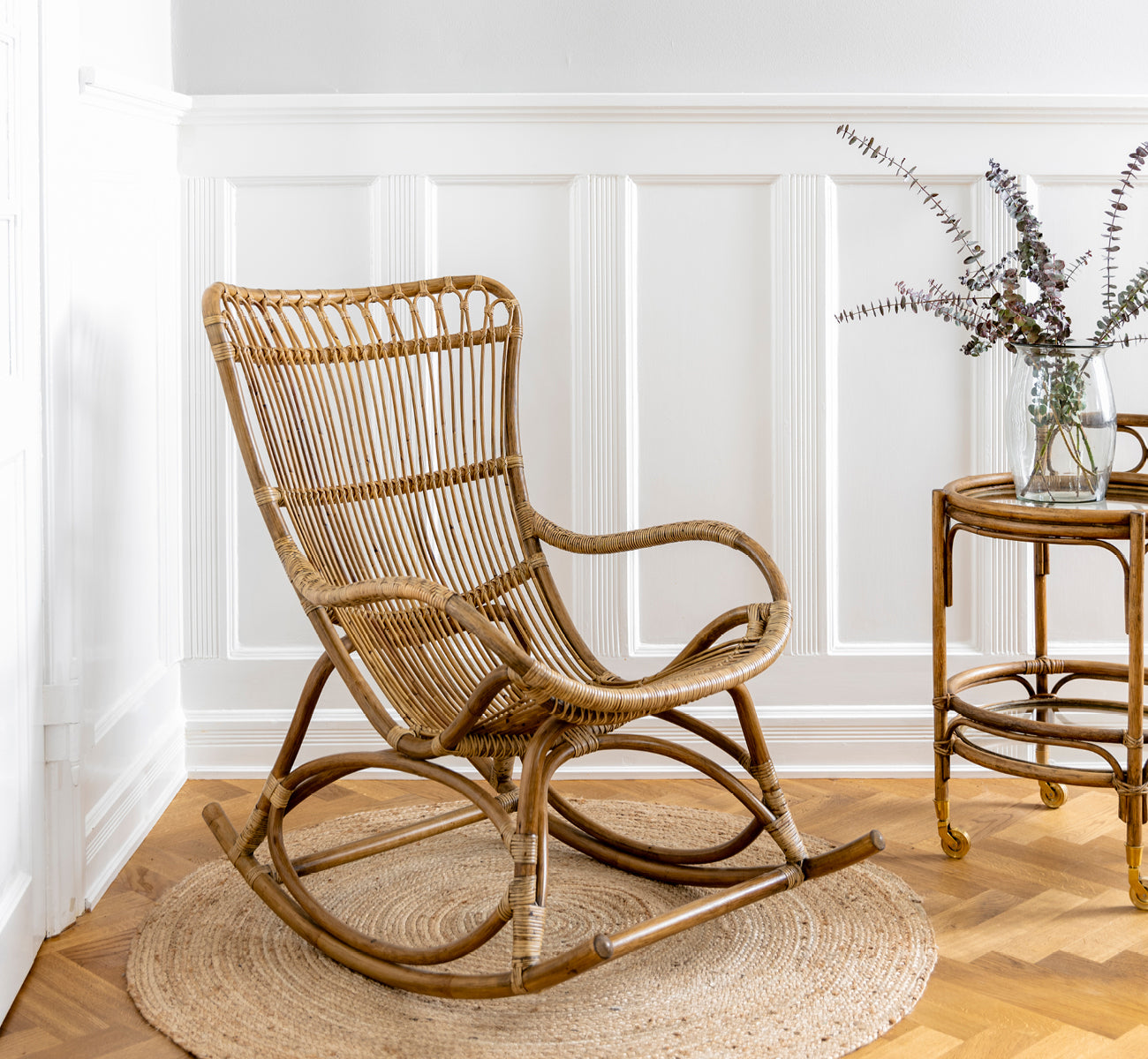 Esta es una imagen de estilo de vida de Monet Rocking Chair i antique rattan. En una habitación con papel tapiz de mariposas y una mesa auxiliar de ratán con tapa de cristal.
