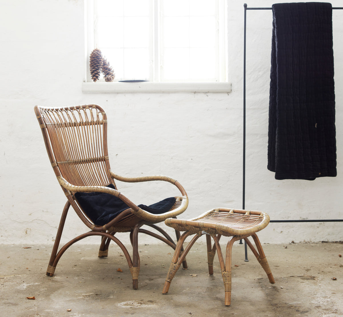 Esta es una imagen de estilo de vida de Monet Lounge Chair i antique rattan. Con reposapiés y mesa auxiliar en un ambiente rústico de granero oscuro.