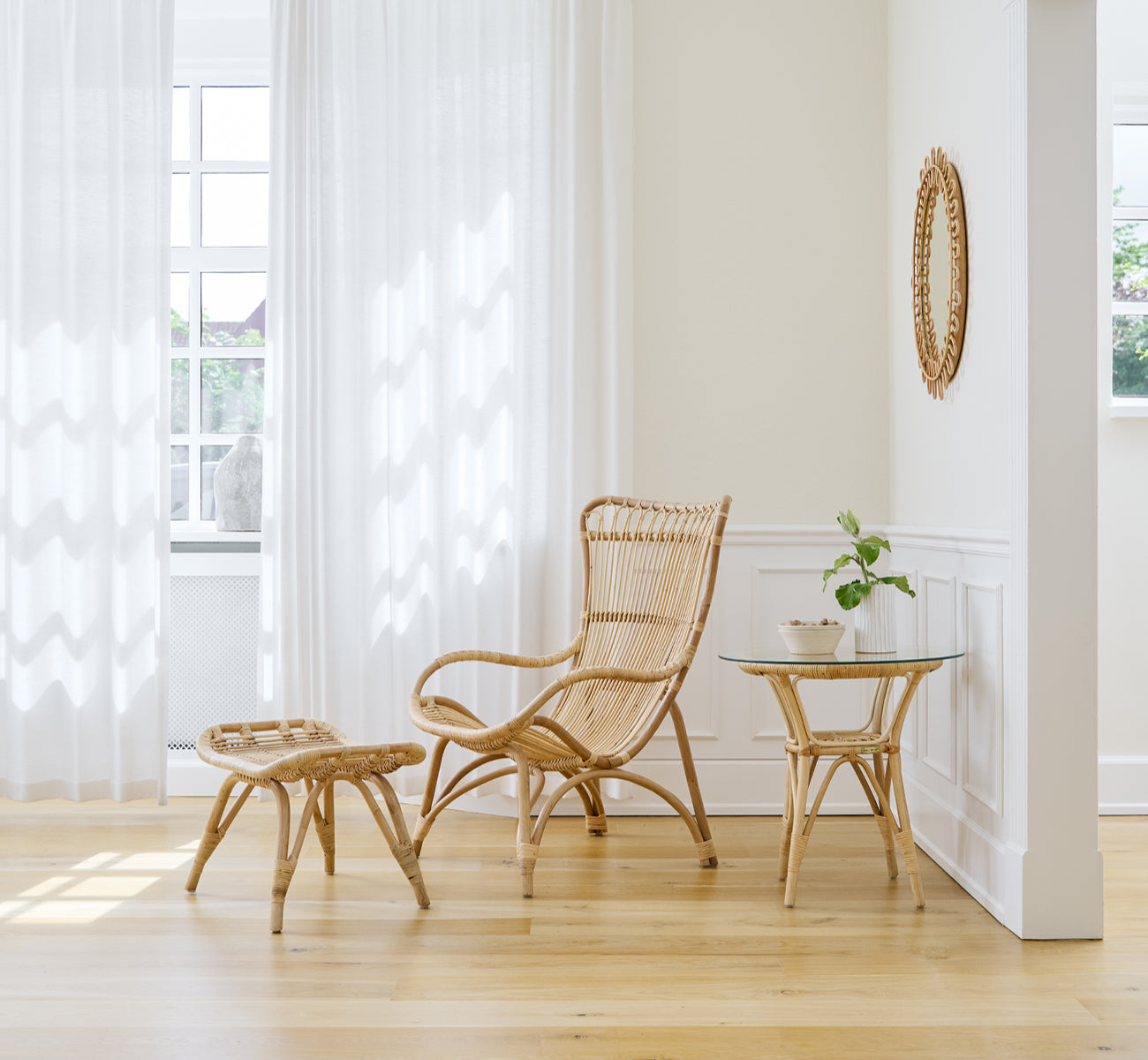 Esta es una imagen de estilo de vida de Monet Lounge Chair i natural rattan. Con reposapiés en una sala luminosa con suelo de roble y cortinas ligeras.