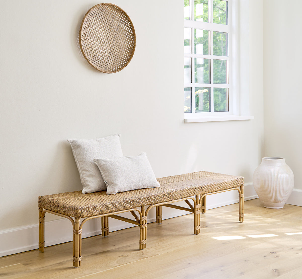 Esta es una imagen de estilo de vida de Luis banco en antique rattan. Estilizado con cojines de lino en una habitación blanca con decoración mural y un jarrón blanco.