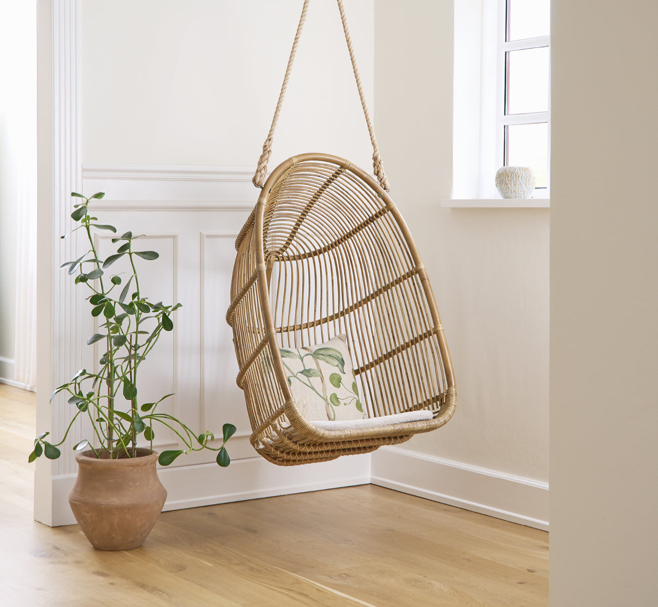 Esta es una imagen de estilo de vida de Renoir silla colgante en antique rattan. Colgada en una sala luminosa con paneles junto a una planta en maceta.
