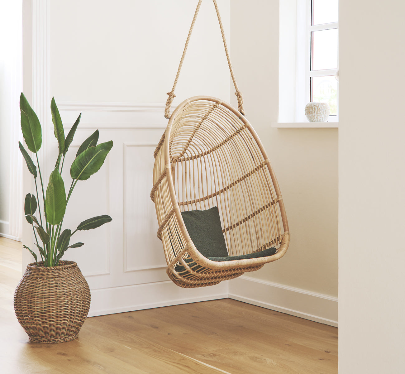 Esta es una imagen de estilo de vida de Renoir silla colgante en natural rattan. Colgada en una entrada luminosa con paneles junto a una planta en maceta.