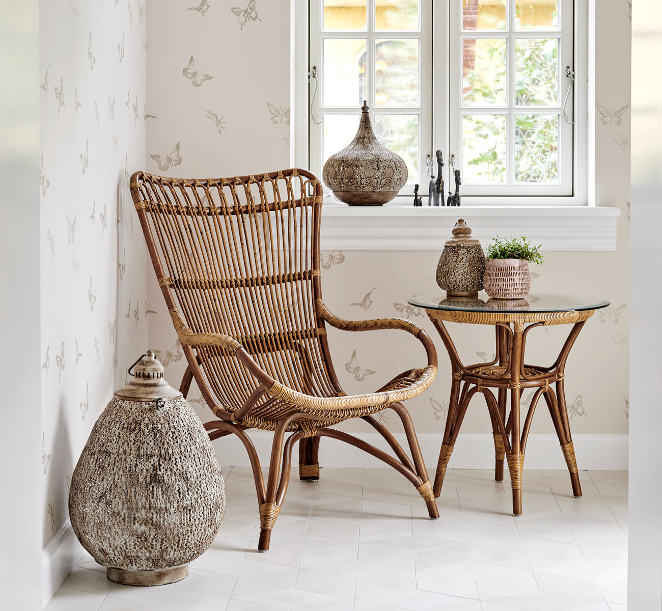 Esta es una imagen de estilo de vida de Tony mesa auxiliar en antique rattan. Colocada en un rincón empapelado con mariposas con linterna y decoración.