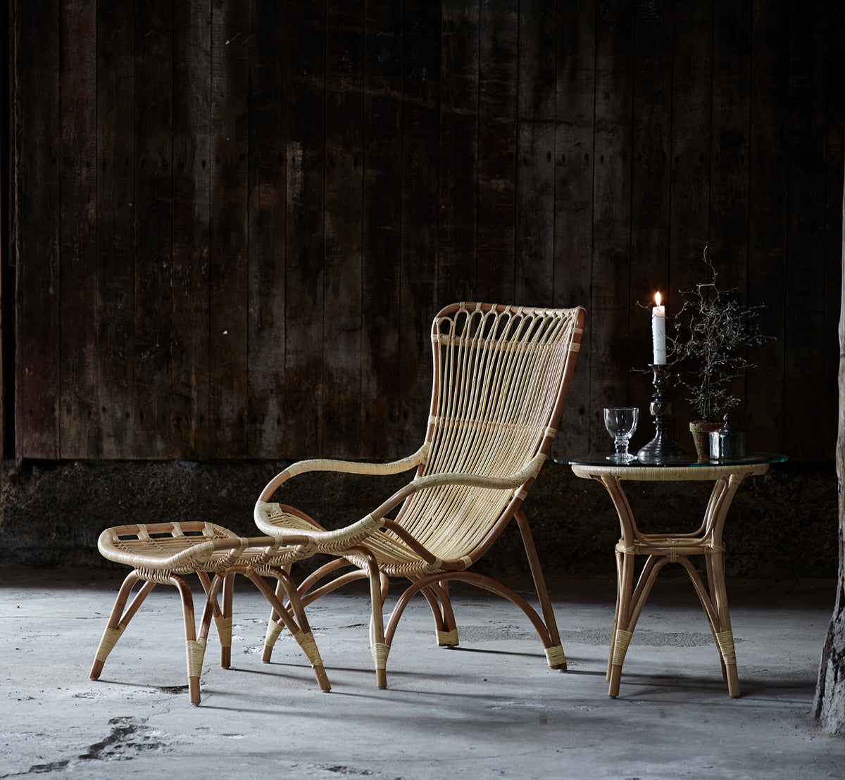 Esta es una imagen de estilo de vida de Tony mesa auxiliar en natural rattan. Paired with a high-back lounge chair against a dark wood wall.
