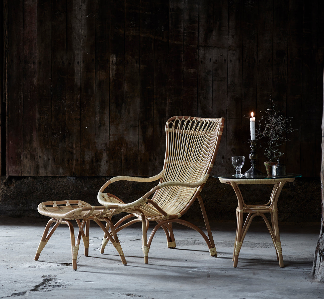 Esta es una imagen de estilo de vida de Tony mesa auxiliar en natural rattan. Paired with a high-back lounge chair against a dark wood wall.