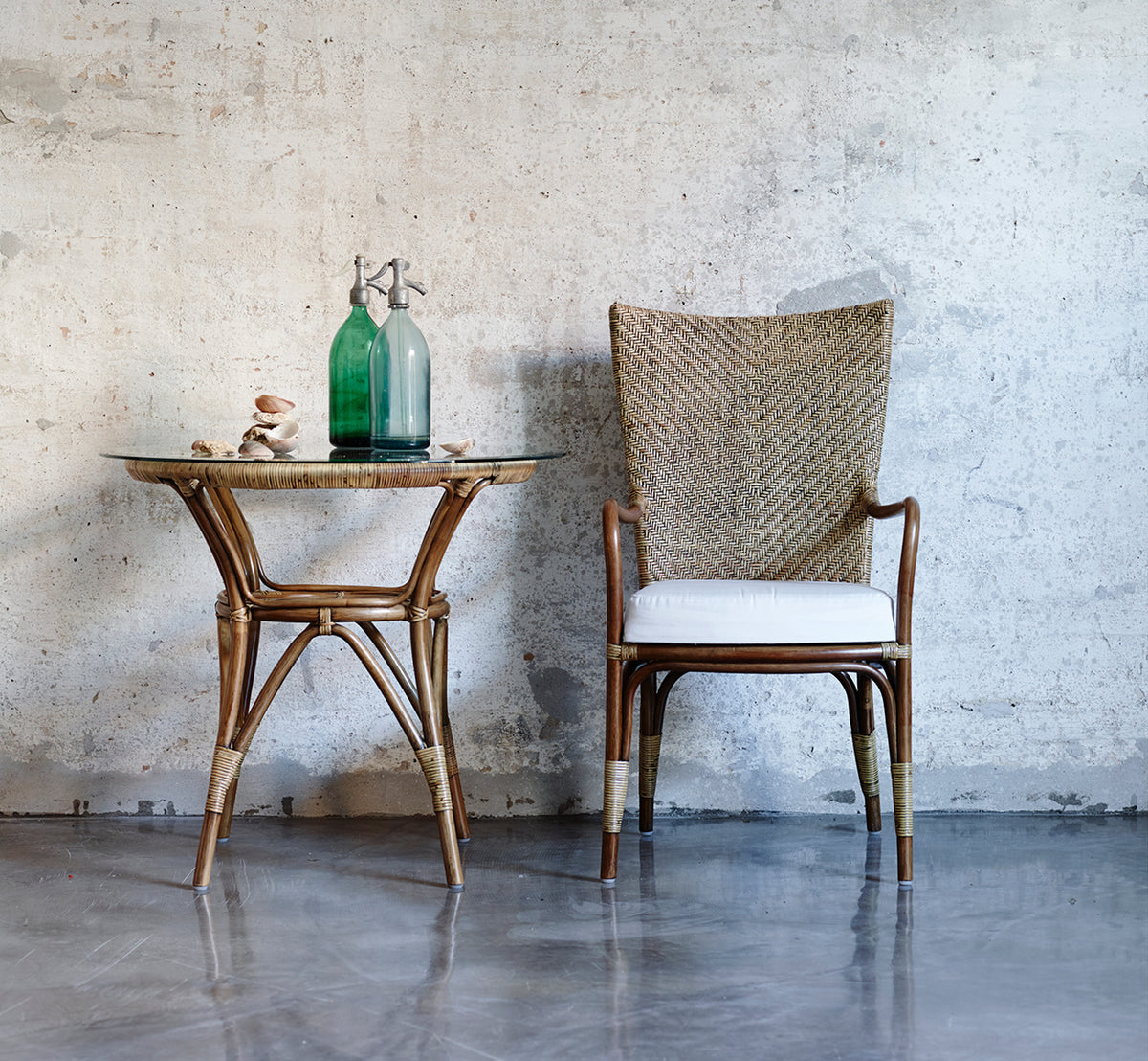 Esta es una imagen de estilo de vida de Tony Café Table en antique rattan. Combinada con una silla de respaldo alto contra una pared de hormigón visto.