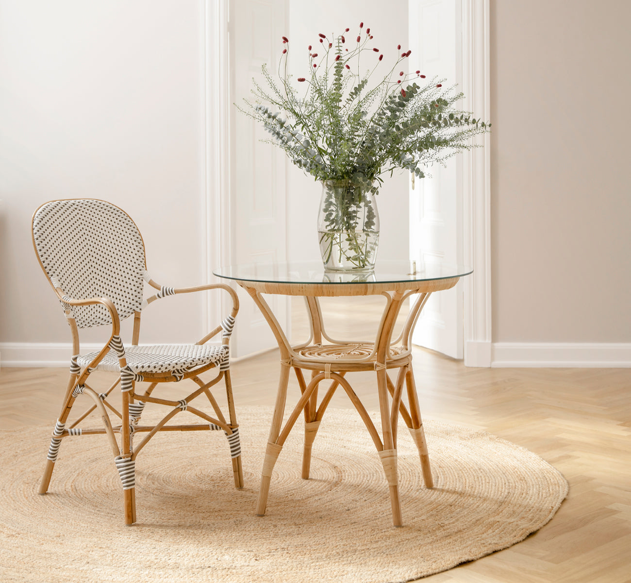 Esta es una imagen de estilo de vida de Tony Café Table en natural rattan. Combinada con dos sillas sobre una alfombra redonda de yute en un umbral luminoso.