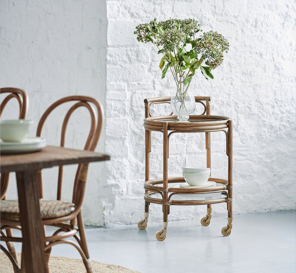 Esta es una imagen de estilo de vida de Romeo carrito en antique rattan. Estilizada con hortensias y cerámica contra una pared de ladrillo blanco.