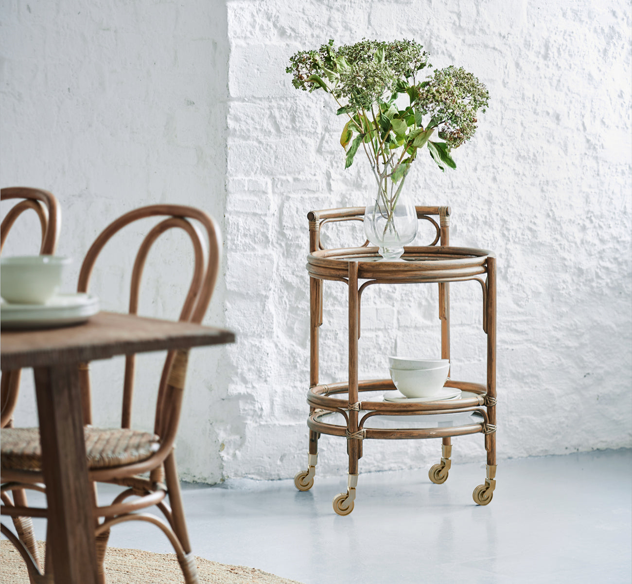 Esta es una imagen de estilo de vida de Romeo carrito en antique rattan. Estilizada con hortensias y cerámica contra una pared de ladrillo blanco.