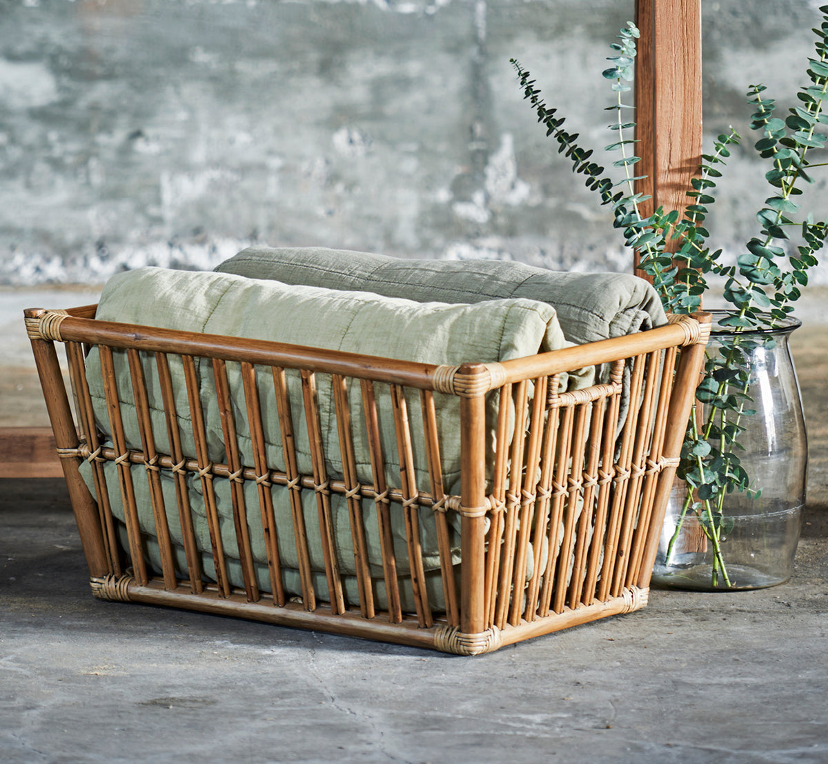 Esta es una imagen de estilo de vida de Marche cesta en antique rattan. Estilizada con mantas de lino y eucalipto en una sala con pared de hormigón.