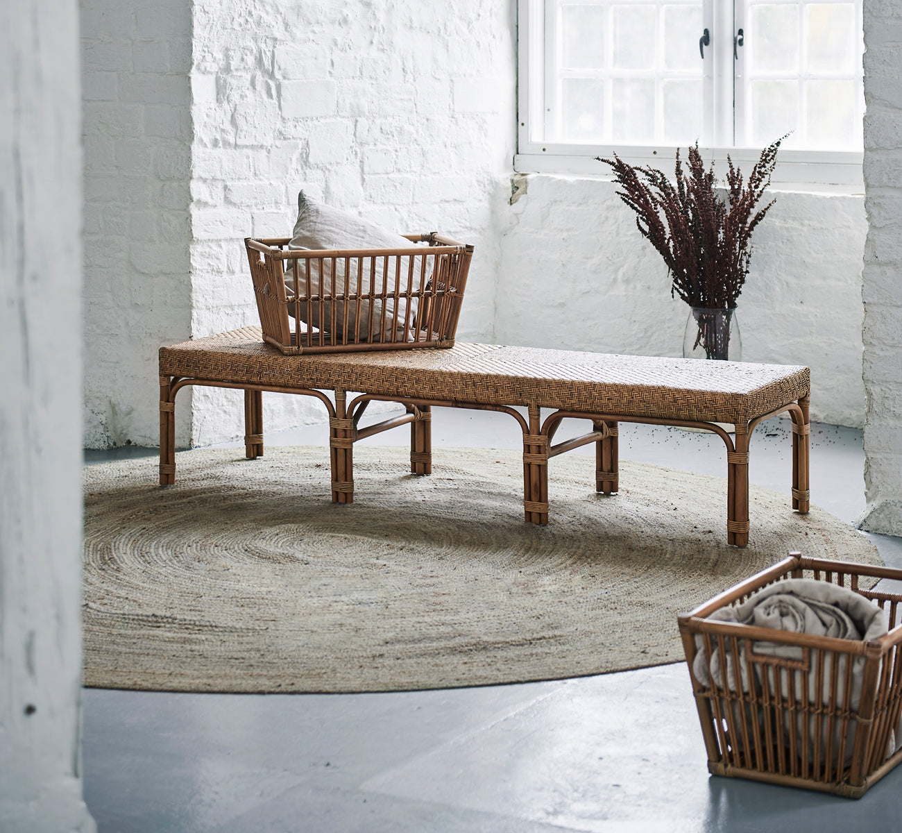 Esta es una imagen de estilo de vida de Marche cesta en antique rattan. Colocada sobre un banco con cojines en un interior de ladrillo blanco con hierbas secas.
