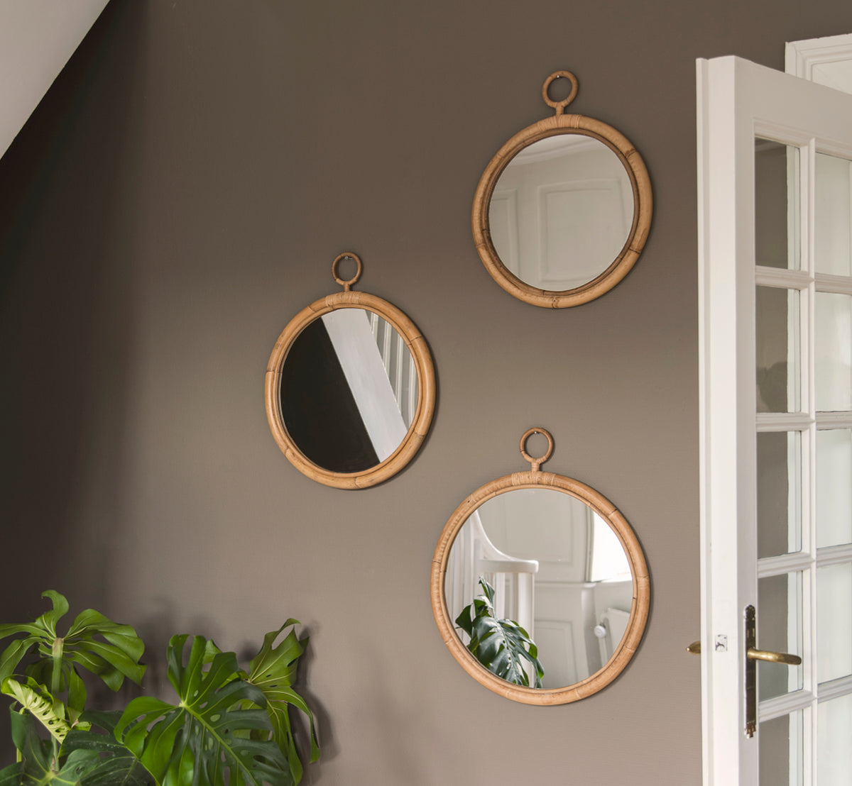Esta es una imagen de estilo de vida de Ella espejo Ø45cm en natural skin on rattan, en una pared color taupé en un pasillo con una planta monstera y.