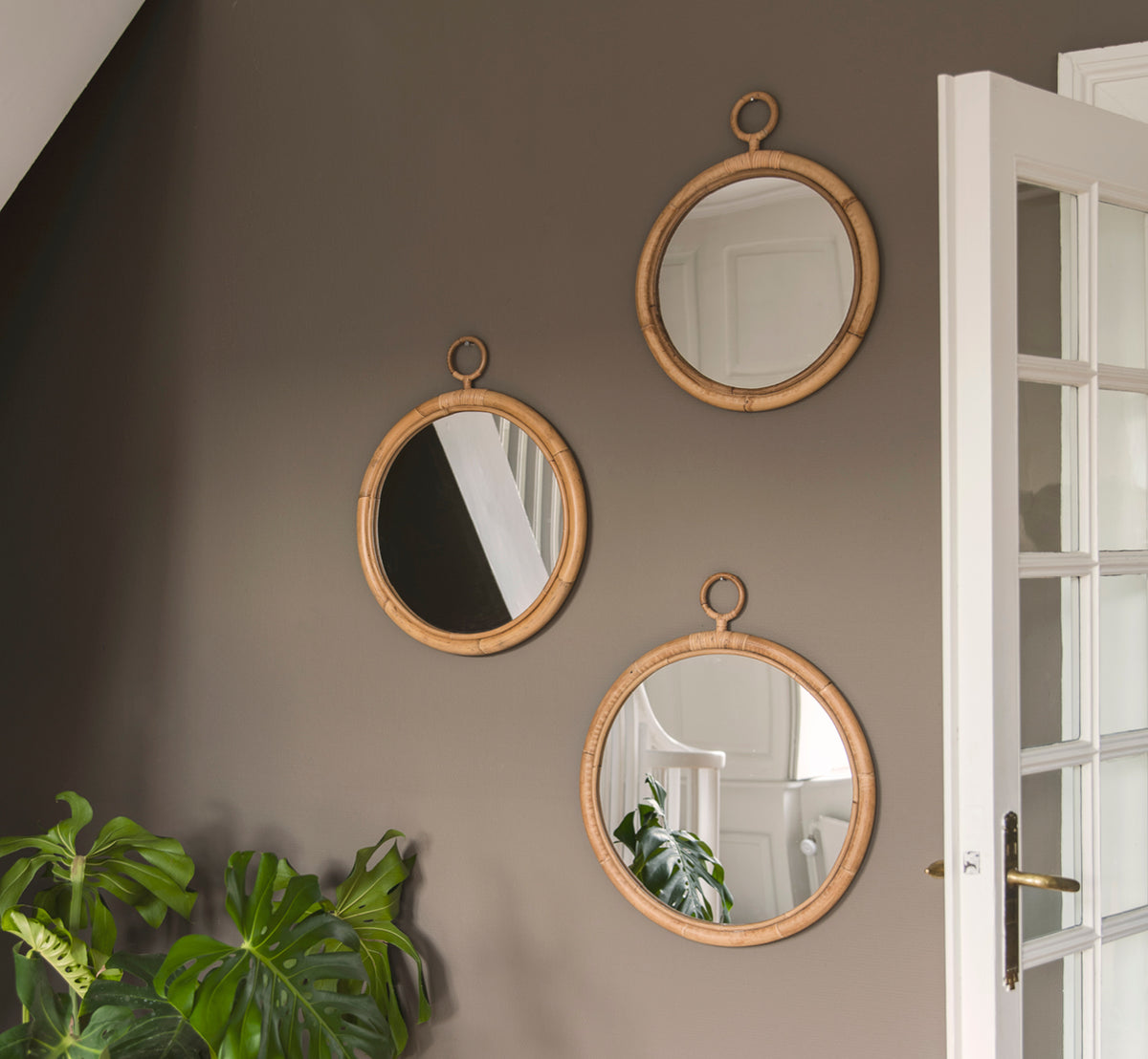 Esta es una imagen de estilo de vida de Ella espejo Ø50cm en natural skin on rattan, en una pared color taupé en un pasillo con una planta monstera y.