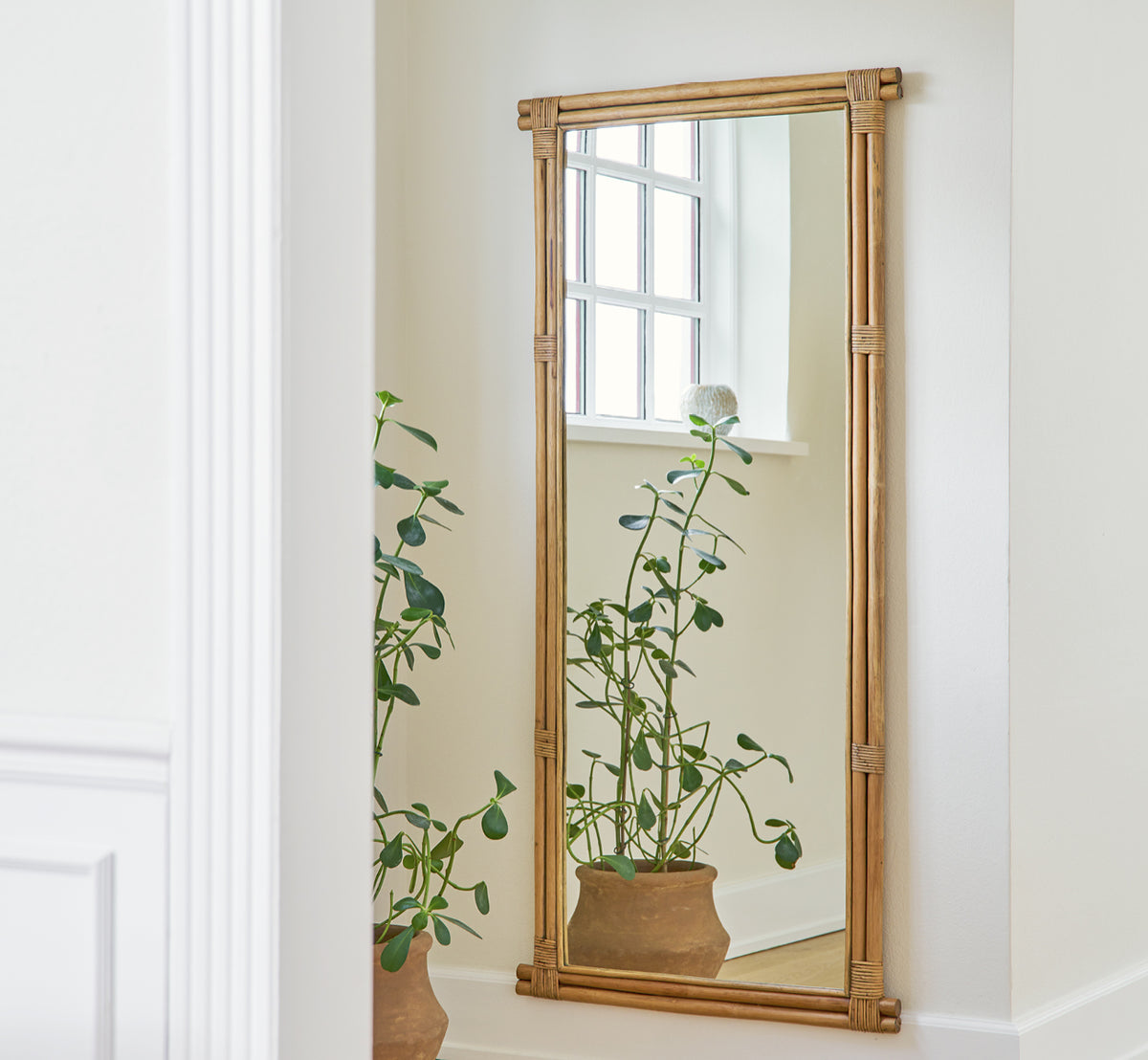 Esta es una imagen de estilo de vida de Rasmus Mirror 172 cm en antique rattan. Montado en una pared blanca sobre una planta en maceta en un pasillo luminoso.