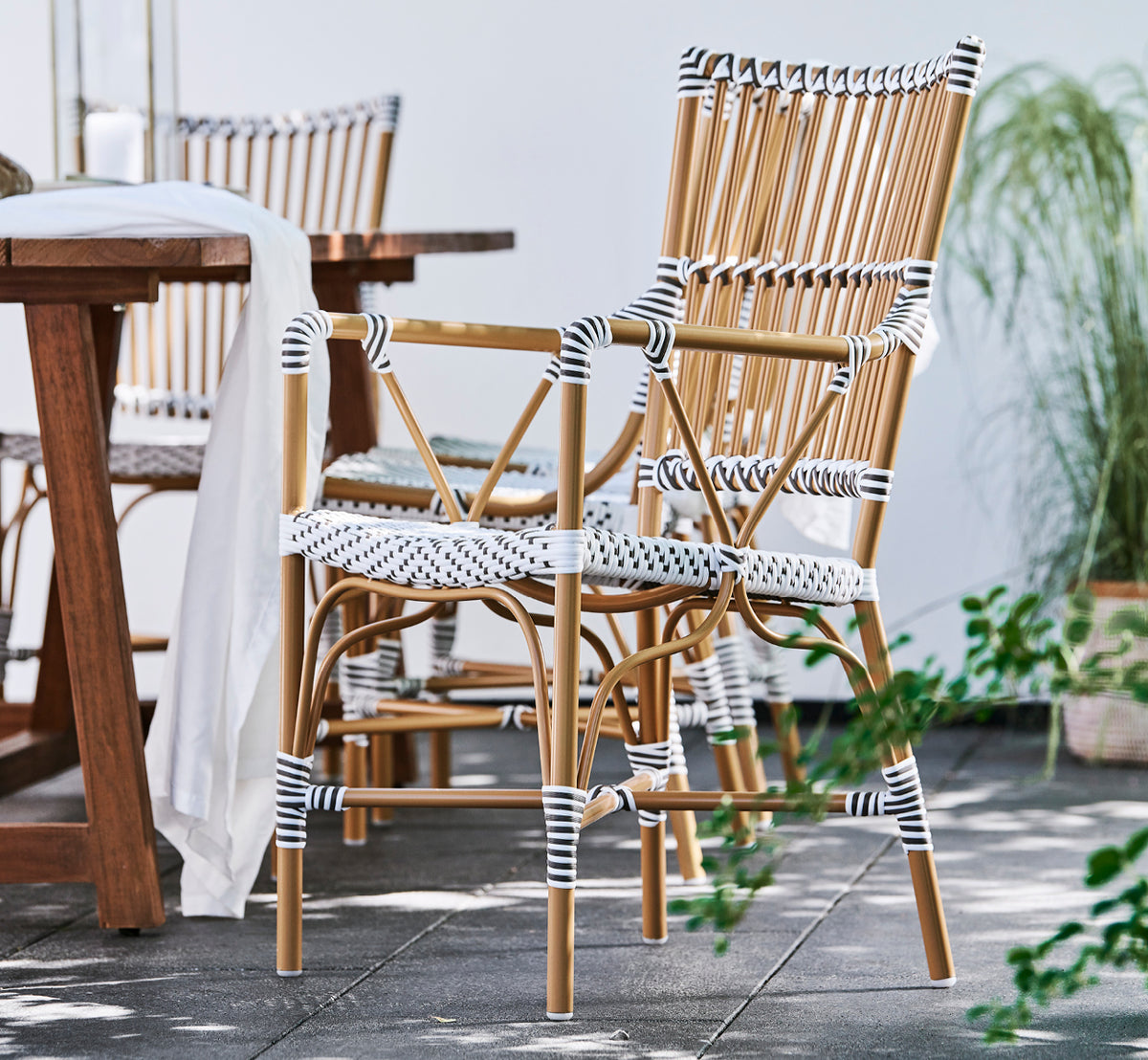 Esta es una imagen de estilo de vida de Monique Exterior Dining Armchair i white w cappuccino dot ArtFibre. En una terraza soleada con palmeras.