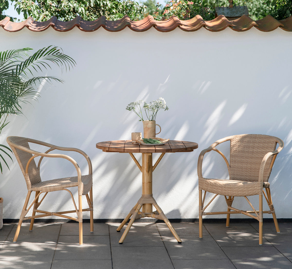 Esta es una imagen de estilo de vida de Madeleine silla de comedor exterior en almond aluminium frame natural ArtFibre. Ambiente de bistró en patio con mesa redonda de teca.