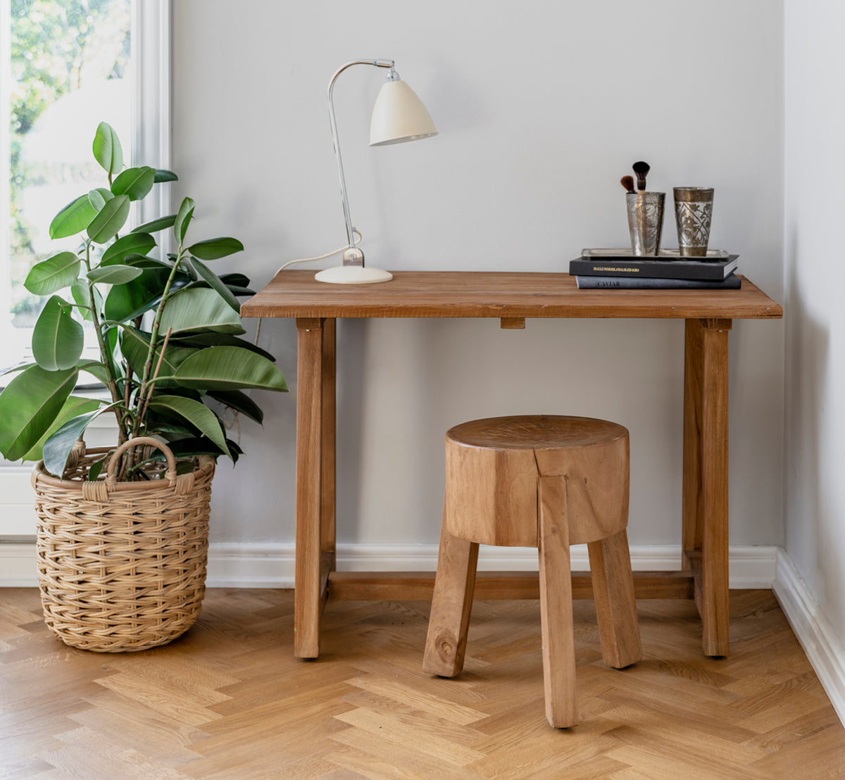 Esta es una imagen de estilo de vida de Roger taburete en teak indoor. Colocada junto a un escritorio de madera con lámpara y planta en un estudio luminoso con suelo en espiga.