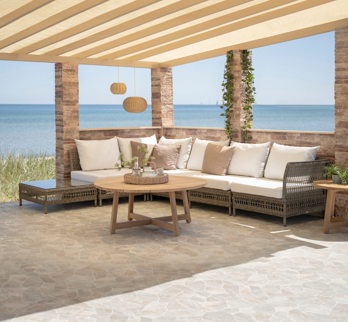 Esta es una imagen de estilo de vida de Mona sofá modular exterior-Pouf Module w. Glass en antique grey artfibre aluminium. Bajo una pérgola junto al mar.