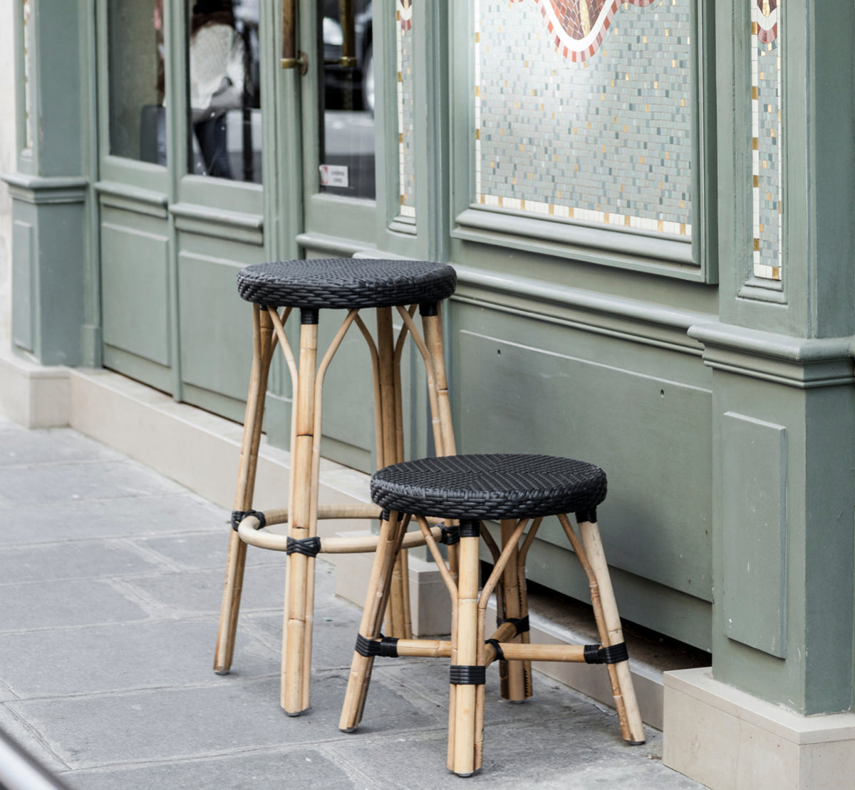 Esta es una imagen de estilo de vida de Simone taburete alto en black ArtFibre Skin on Rattan frame. Frente a una fachada de café parisino.