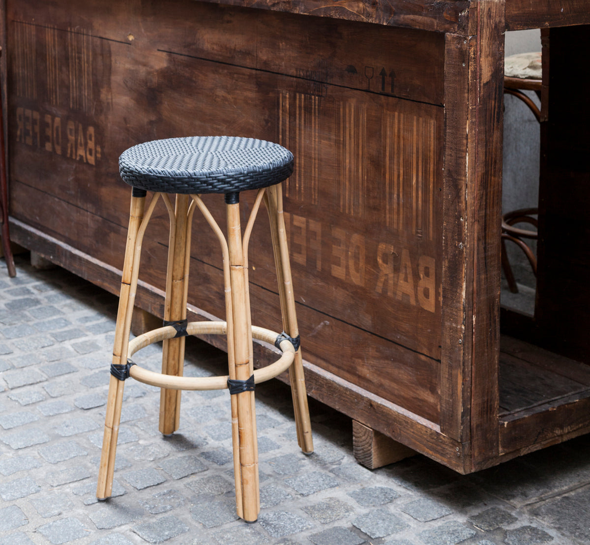 Esta es una imagen de estilo de vida de Simone taburete de bar en black ArtFibre Skin on Rattan frame. Junto a un bar de madera sobre adoquines.